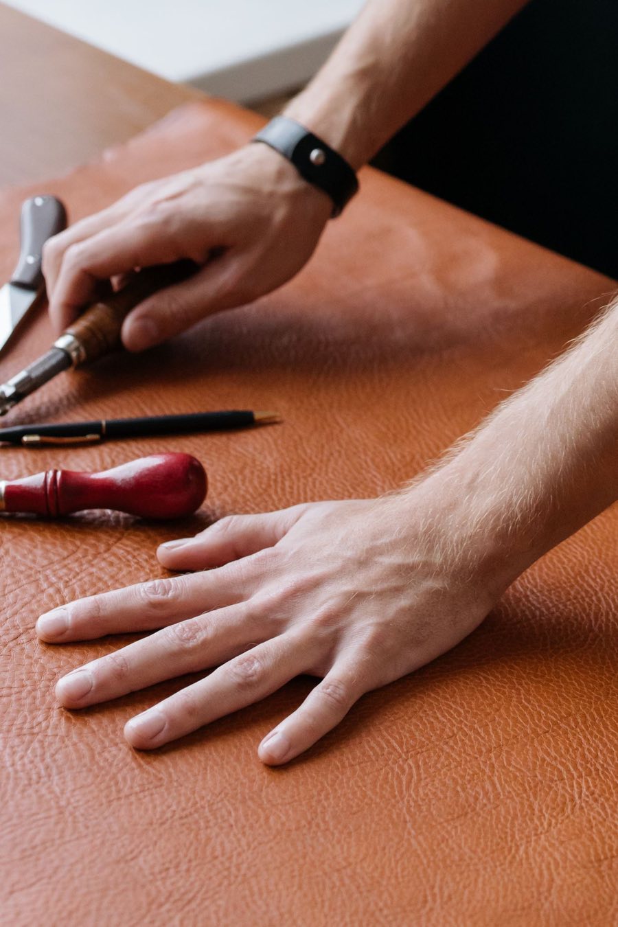 Hands and hand tools working a piece of rich tan leather on a bench